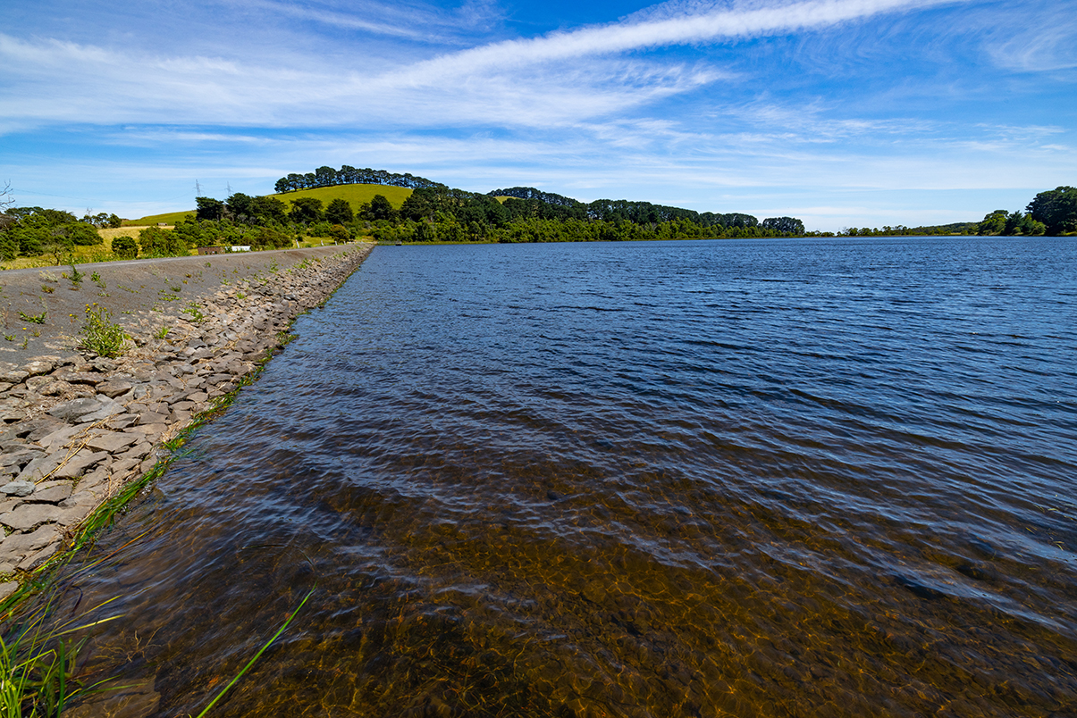 Rippling water in a water reservoir with an embankment on the left and greenery in the background.