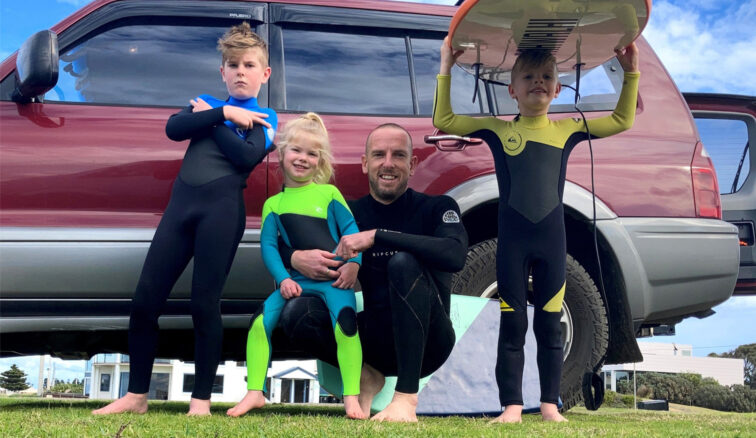Family-friendly, Linton Hall and children, crouching wearing wetsuits, holding surfboard