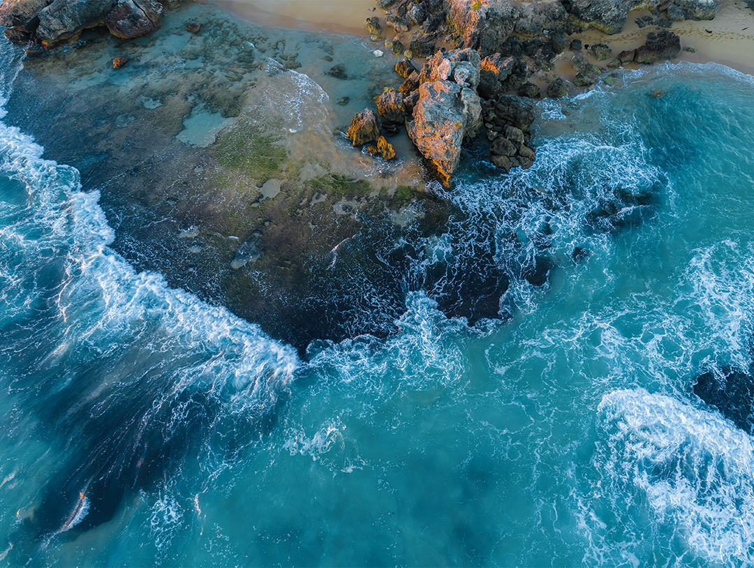 A drone image of small waves rolling over sandstone reef onto shore