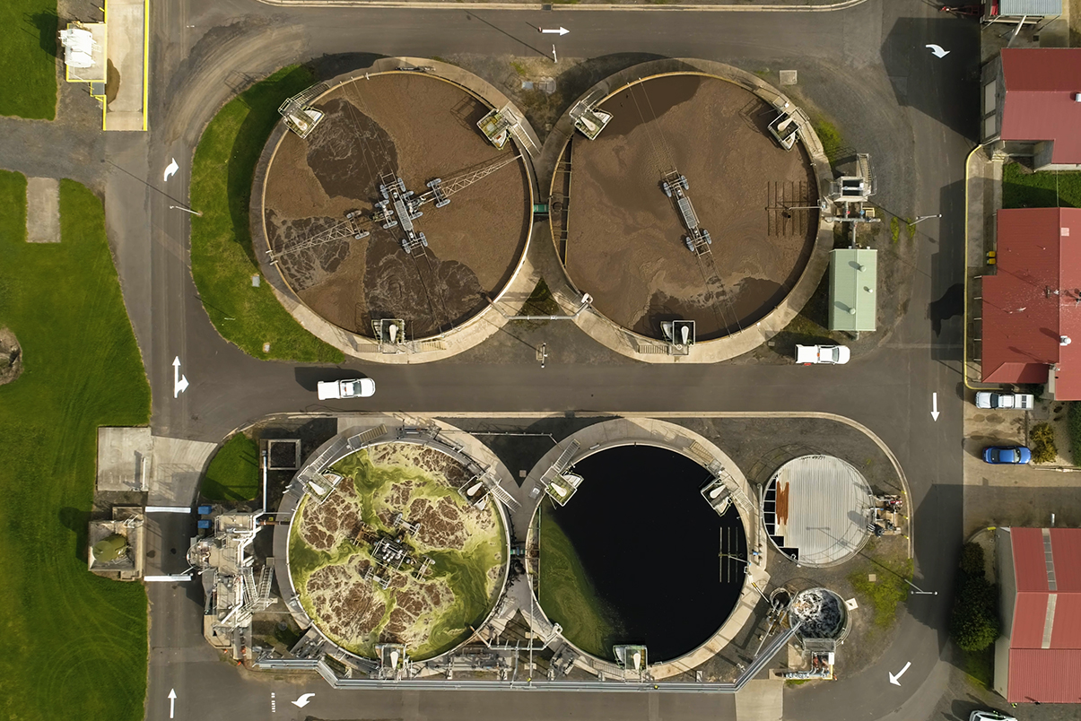 From overhead we see four sewage treatment tanks in operation surround by grass on the left and red roofed buildings on the right.