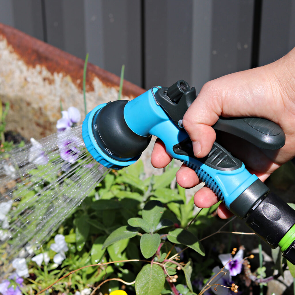 Hand holding a light blue trigger nozzle while spraying water on light purple flowers.