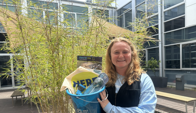 Smiling Wannon Water employee Jess holds a bucket with a soil water meter, trigger nozzle and tote bag. She's standing in front of a plant on a sunny day.